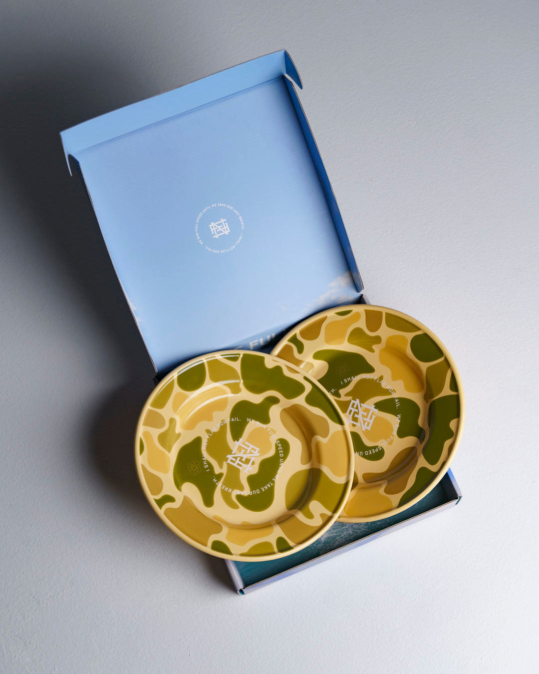 An open box revealing 2 camo-patterned enamel plates, against white studio background