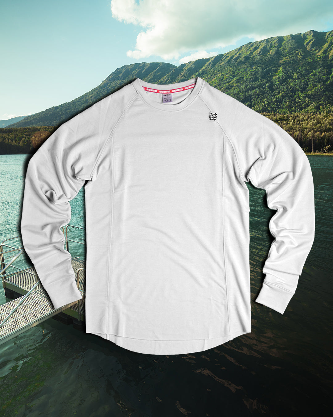 White long-sleeve shirt with brand logos on a dock with mountains and water in the background