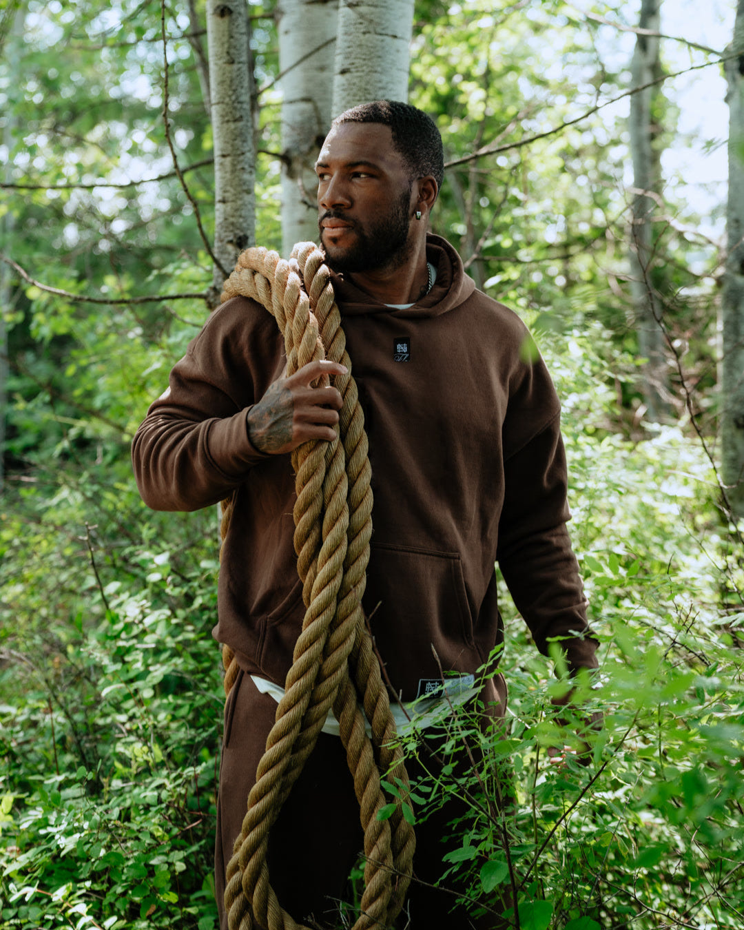 Man wearing brown hoodie and sweatpants holding a large rope in a forest setting