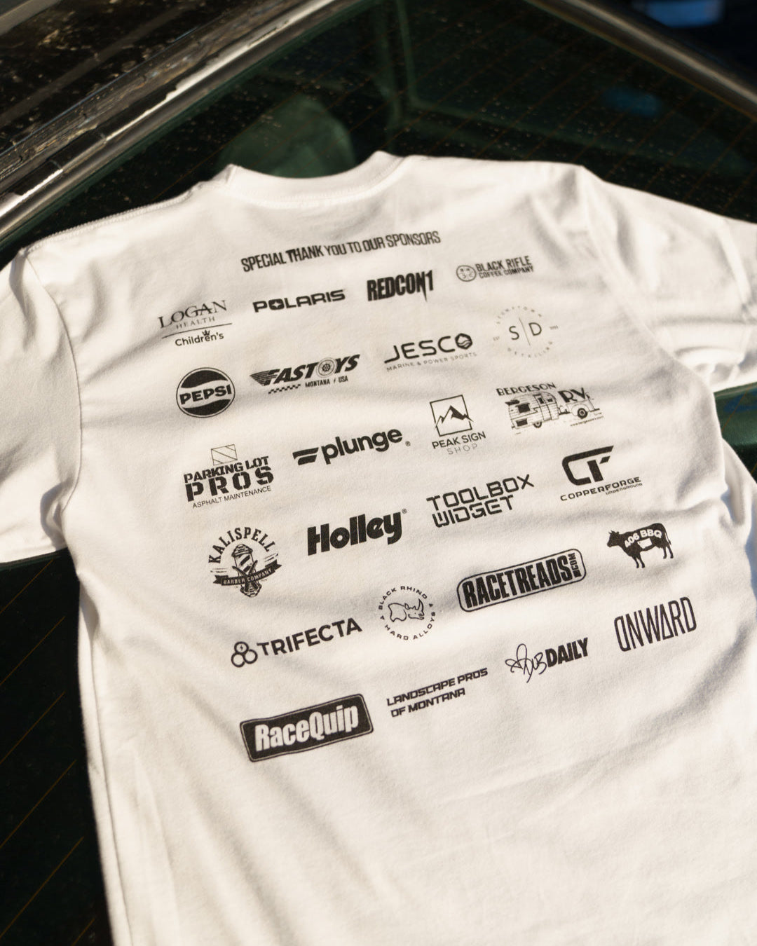 White t-shirt with various brand logos on a car rear wind shield
