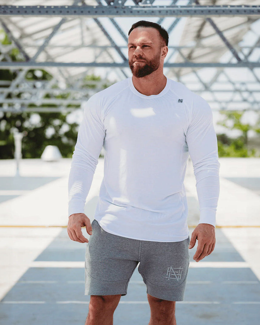 Man wearing a white long-sleeve shirt and gray shorts on a rooftop with greenery in the background