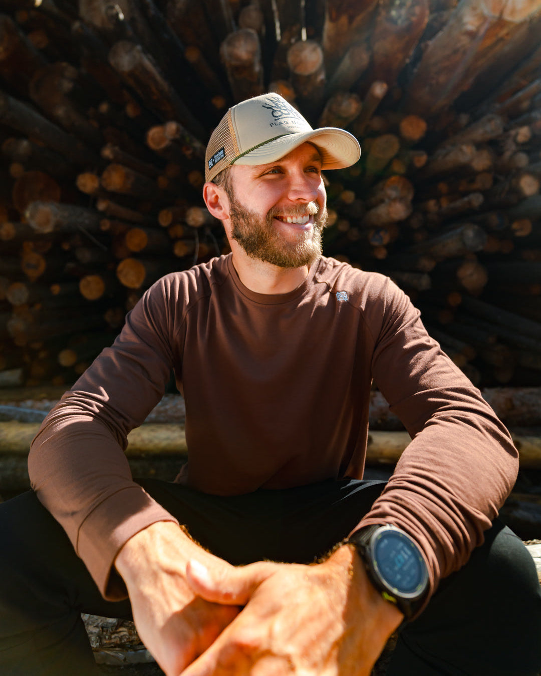 Man sitting in front of a stack of logs wearing a cap and brown long-sleeve shirt.