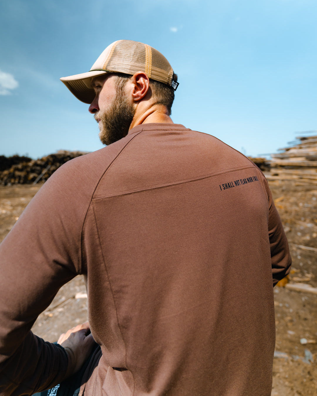 Man wearing a brown jacket with text on the back, standing outdoors with a clear blue sky.