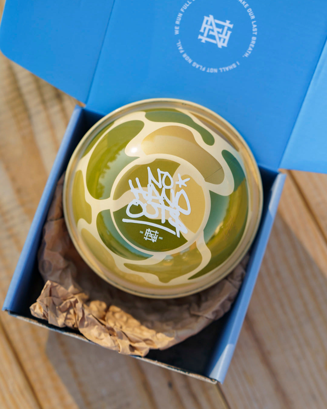 Ceramic bowl with green and brown camo design facing down in a blue box on a wooden picnic table, top down photo.