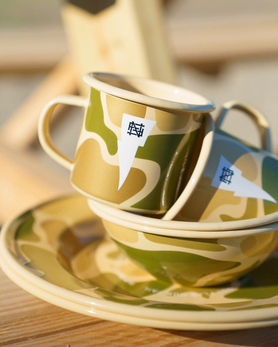 Camo ceramic cup, bowl and plate set stacked on top of each other on a wooden picnic table.