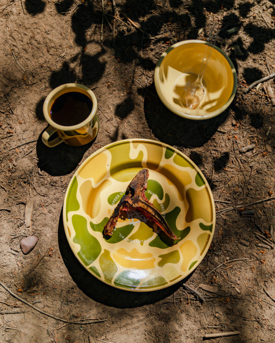 A camo-patterned ceramic mug, bowl and plate with food items; siting on ground