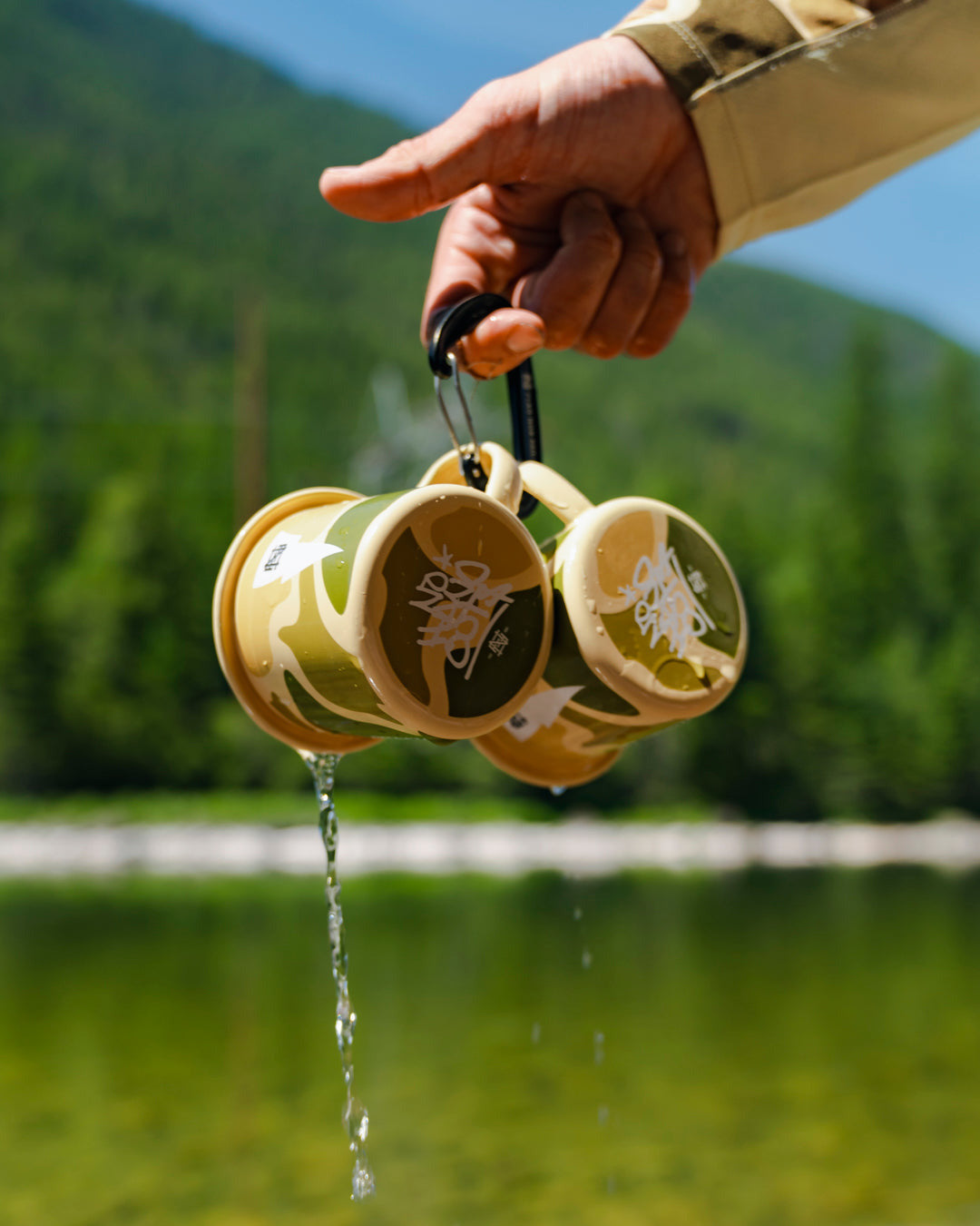 Hand holding a set of two yellow enamel cups over a body of water with mountains in the background.