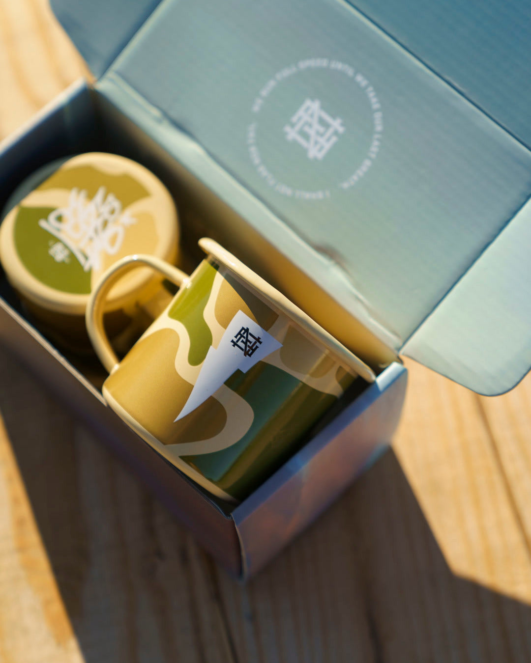 Two camo ceramic mugs with FNF monogram logo in an open box on a wooden picnic table.