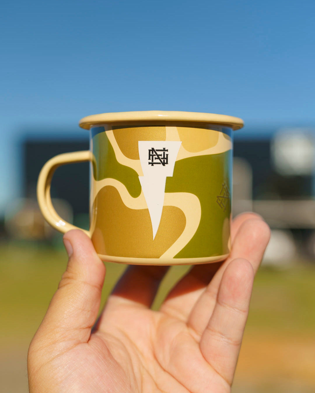 Hand holding a camo ceramic mug with FNF monogram logo against a blurred outdoor background