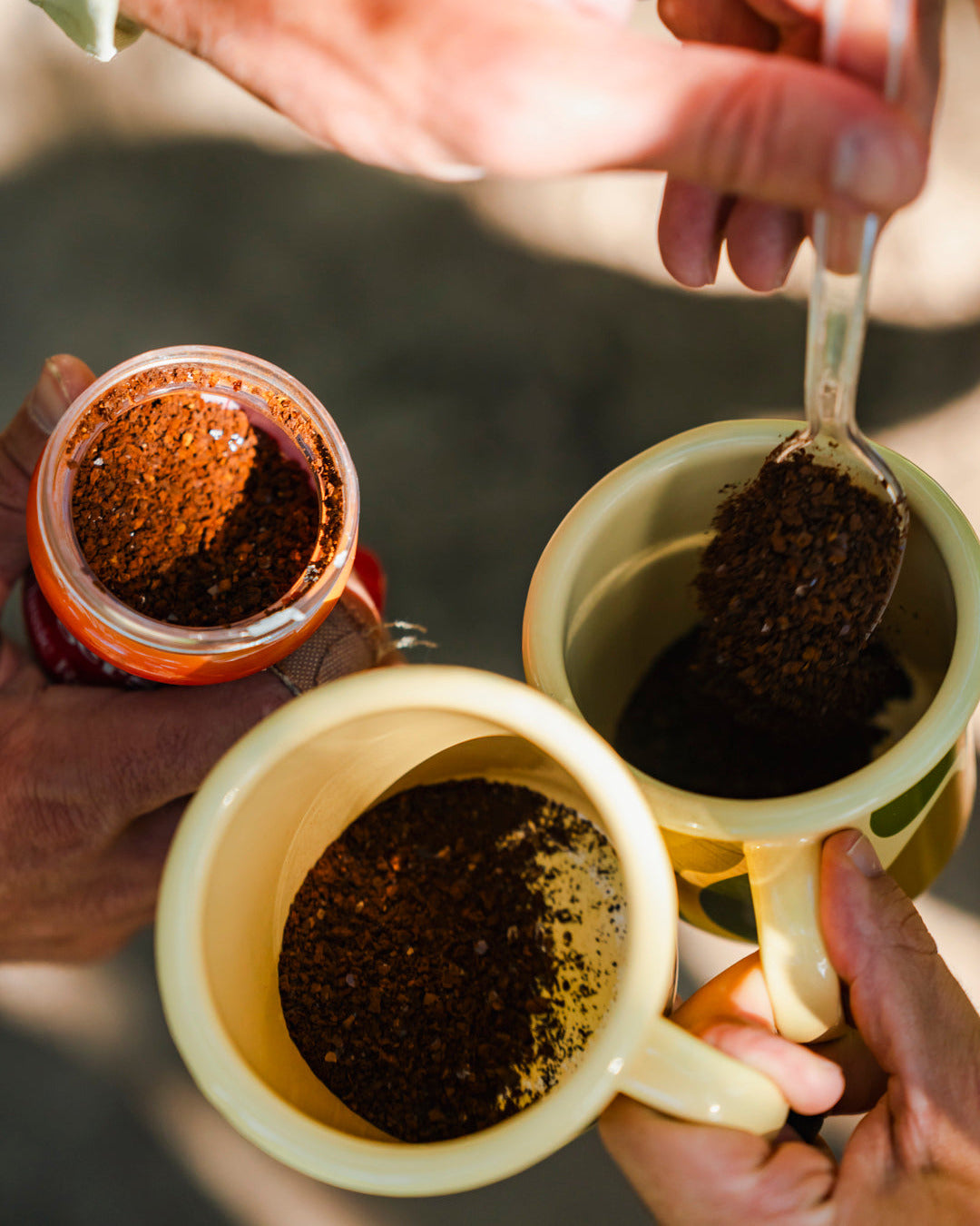 Two ceramic mugs getting filled with coffee grains, held by hands against a blurred background outdoors.