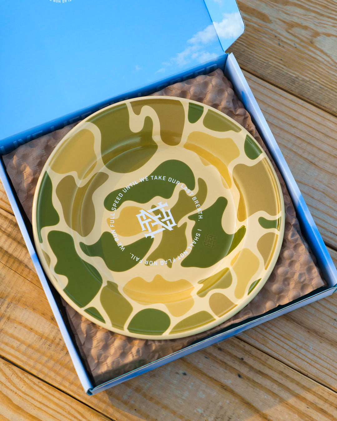 A camo-patterned enamel plate in a blue box on a wooden picnic table