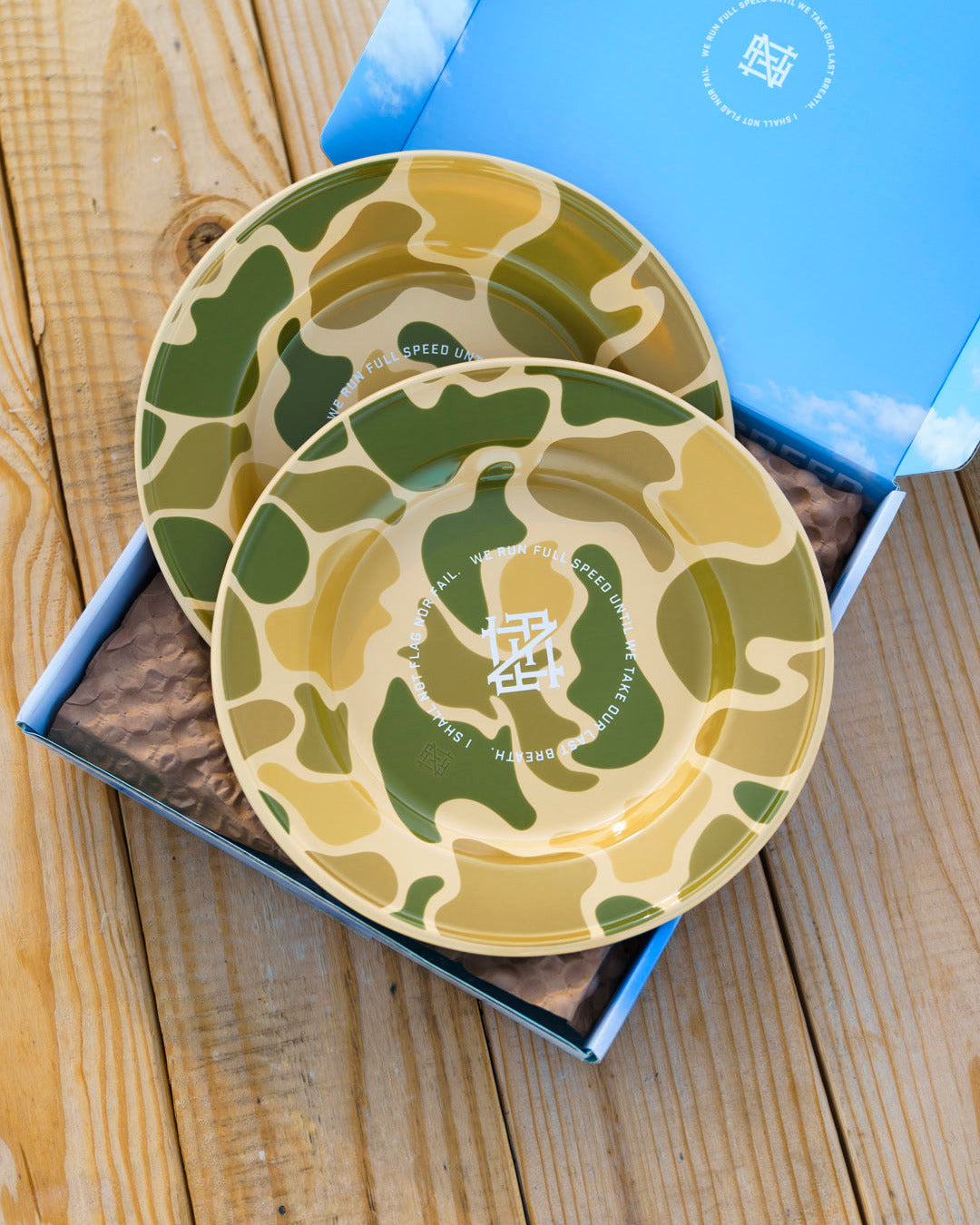 Two camo-patterned enamel plates on a wooden picnic table within a blue box.