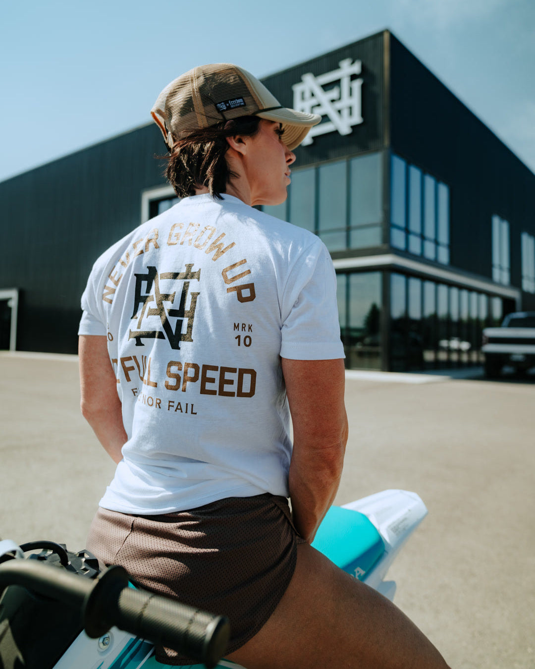 Person wearing a white t-shirt with text and graphics, sitting on a motorcycle in front of a building.