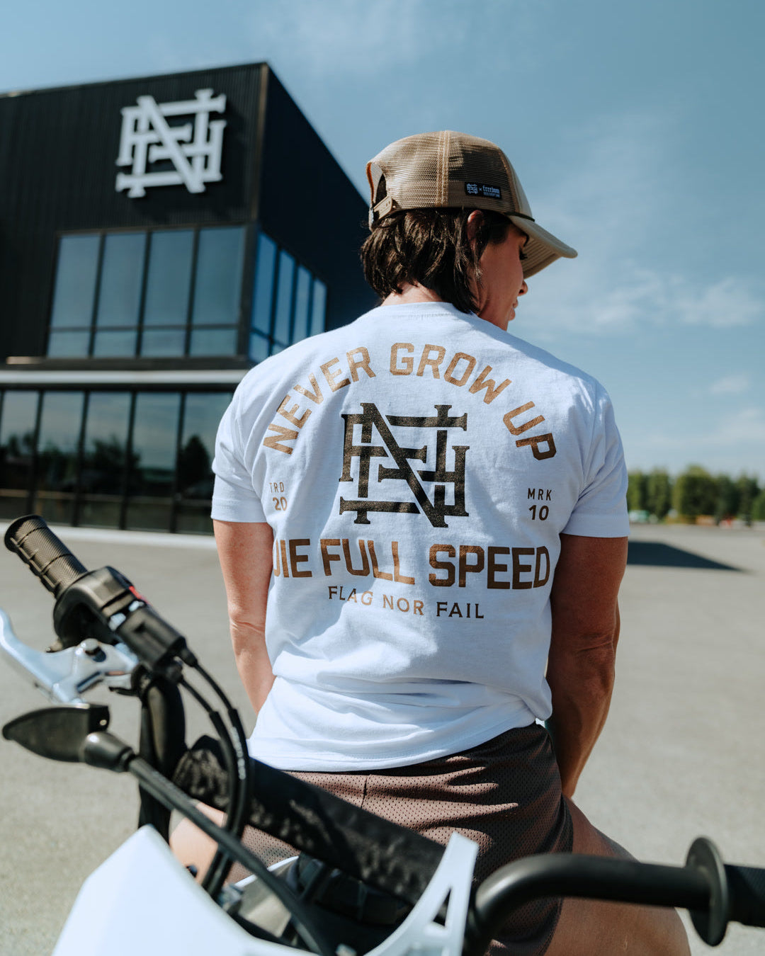 Person wearing a white t-shirt with text and logo, sitting on a motorcycle outdoors.